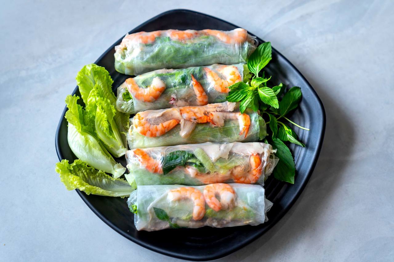 Năm cuốn gỏi cuốn tôm nhìn thấu qua lớp bánh tráng, đặt trên đĩa đen với xà lách và rau húng