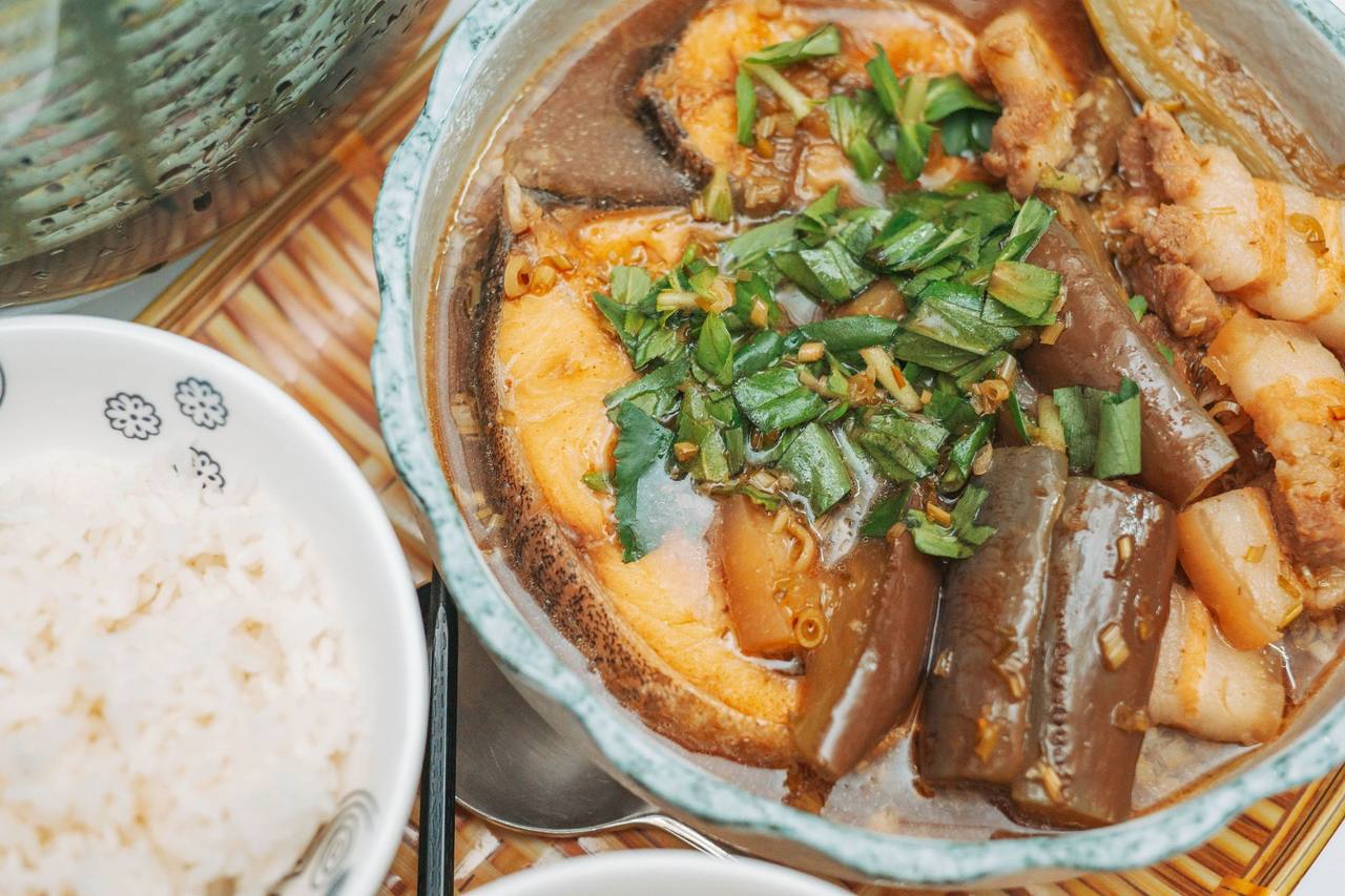 Nồi cá kho cà tím rắc hành lá, kèm chén cơm trắng, nhìn từ trên xuống trên mẹt tre