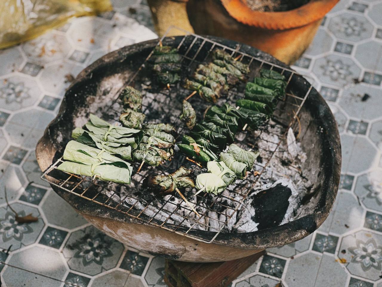 Những cuốn lá lốt nướng trên vỉ sắt đặt trên lò than đất nung, sàn gạch hoa phía dưới