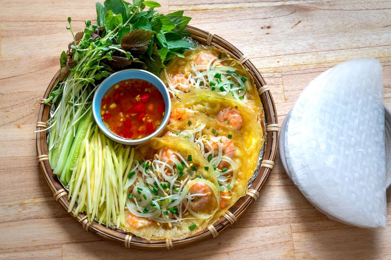 Bánh xèo vàng nghệ nhân tôm, giá, kèm xoài bào sợi, rau thơm, chén nước mắm ớt và miếng bánh tráng, trên mẹt tre