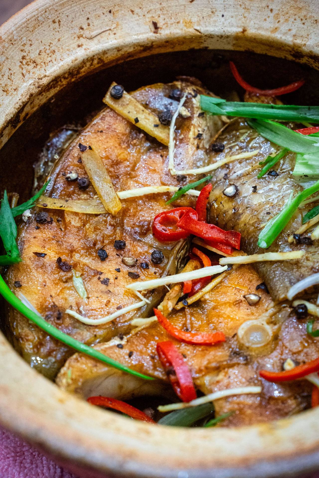 Cá kho nước màu trong nồi đất, rắc hành lá, gừng sợi và ớt đỏ cắt lát