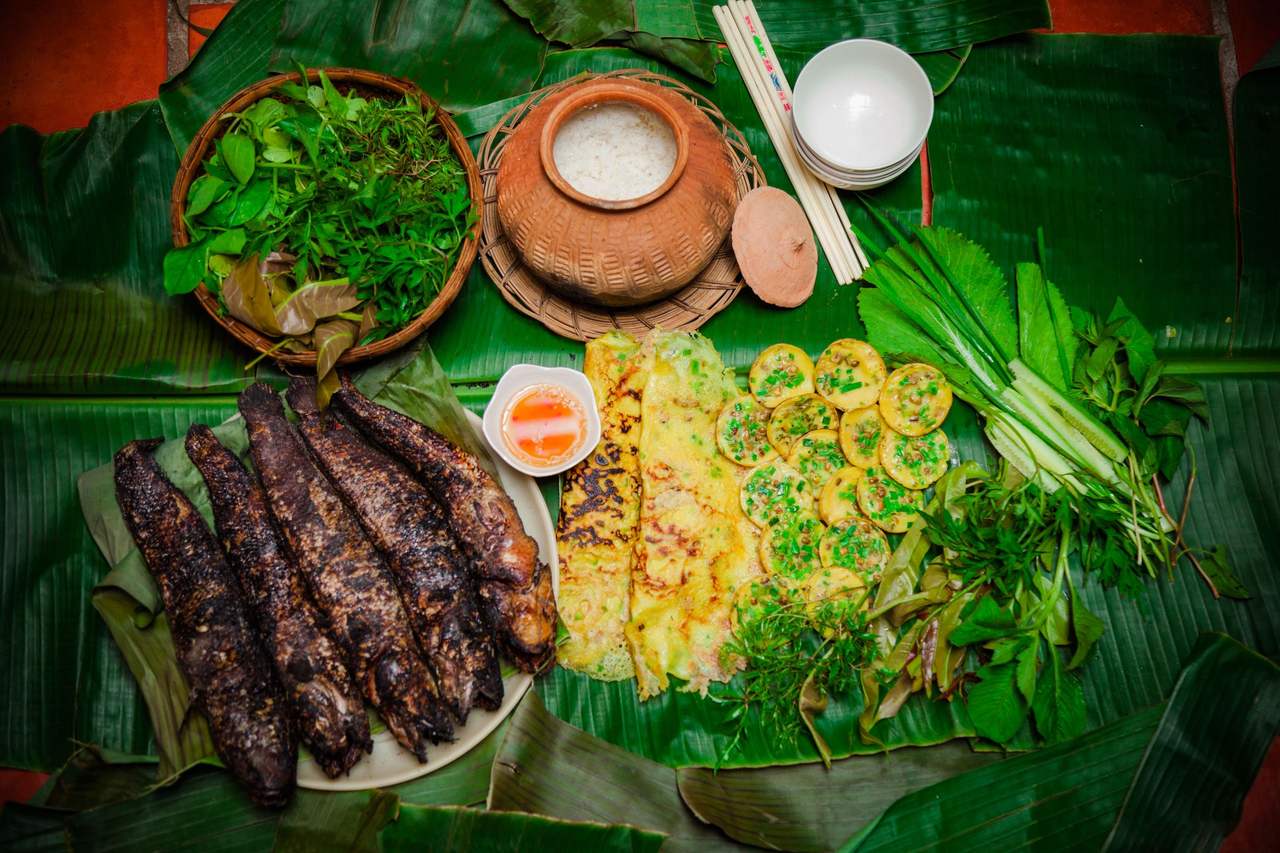 Ba con cá lóc nướng trui cháy sém đặt trên lá chuối, bên cạnh bánh xèo, rổ rau thơm và nồi đất cơm