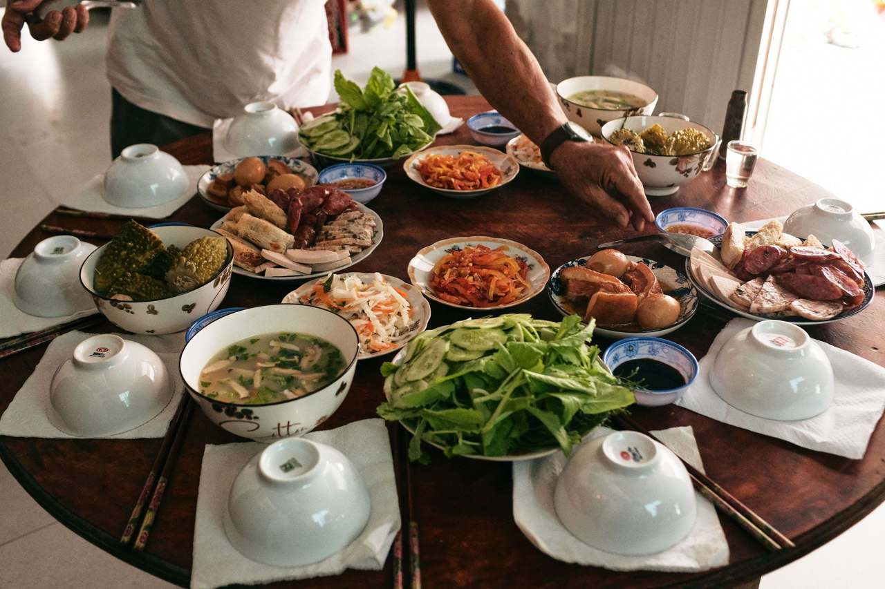 Mâm cơm gia đình trên bàn tròn gỗ: tô canh, rau sống, thịt kho trứng, chả, đồ chua và các chén úp trên khăn giấy, một cánh tay đang gắp món