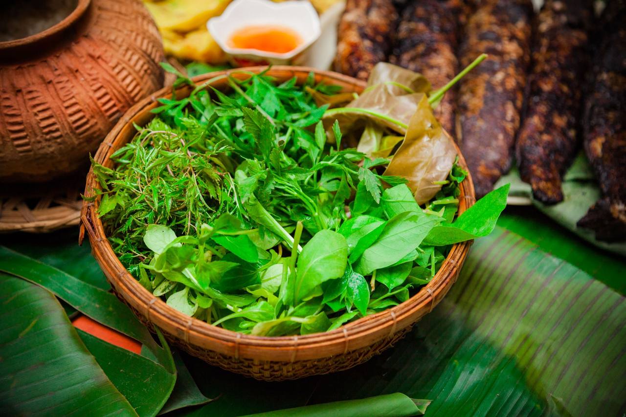Rổ tre đầy rau thơm tươi đặt trên lá chuối, phía sau là nồi đất và cá nướng