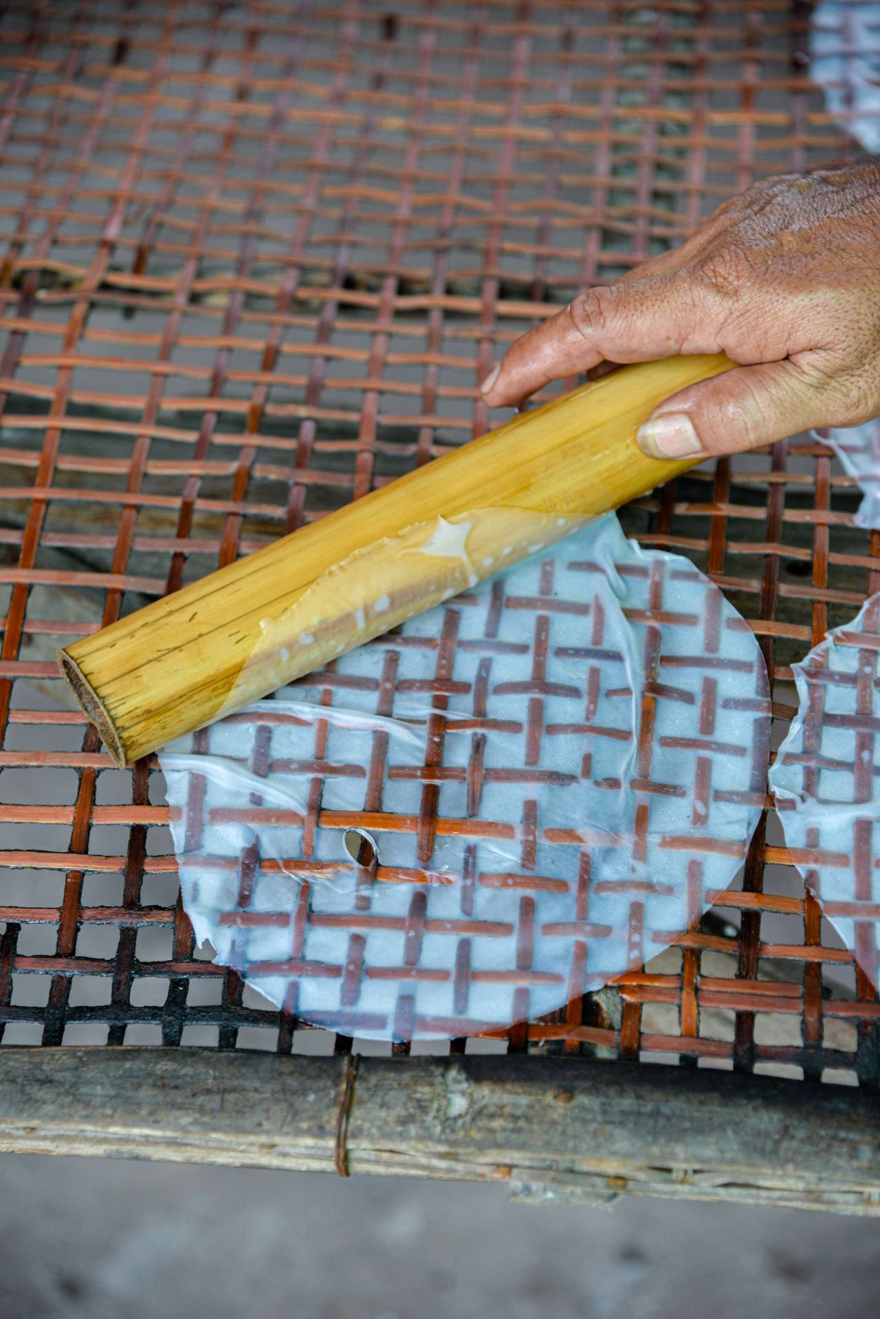 Bàn tay dùng ống tre gỡ tấm bánh tráng mỏng vừa tráng xong trên giàn tre