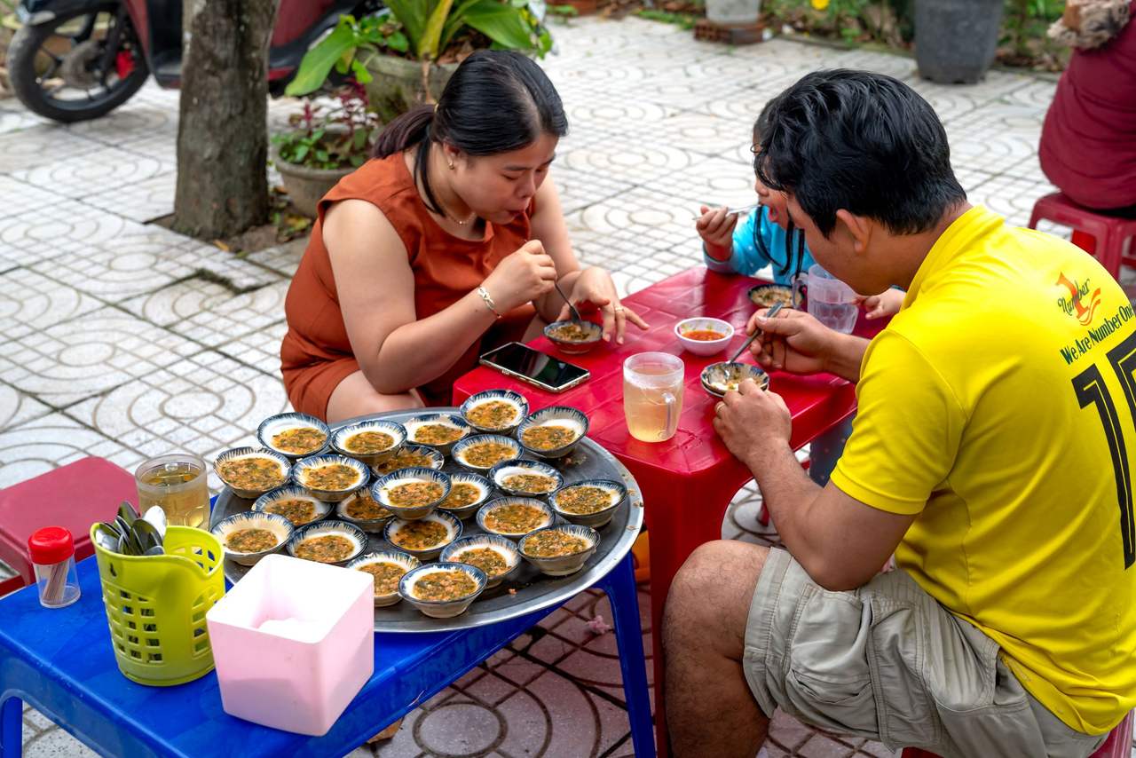 Một gia đình ngồi ghế nhựa thấp quanh khay bánh khọt trên vỉa hè lát gạch, chấm nước mắm