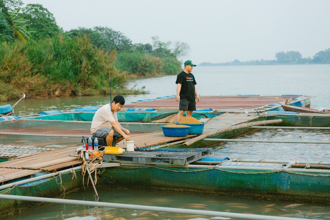 Hai người nuôi cá trên bè nổi bên sông, lồng cá xanh dưới mặt nước và bờ cây phía xa