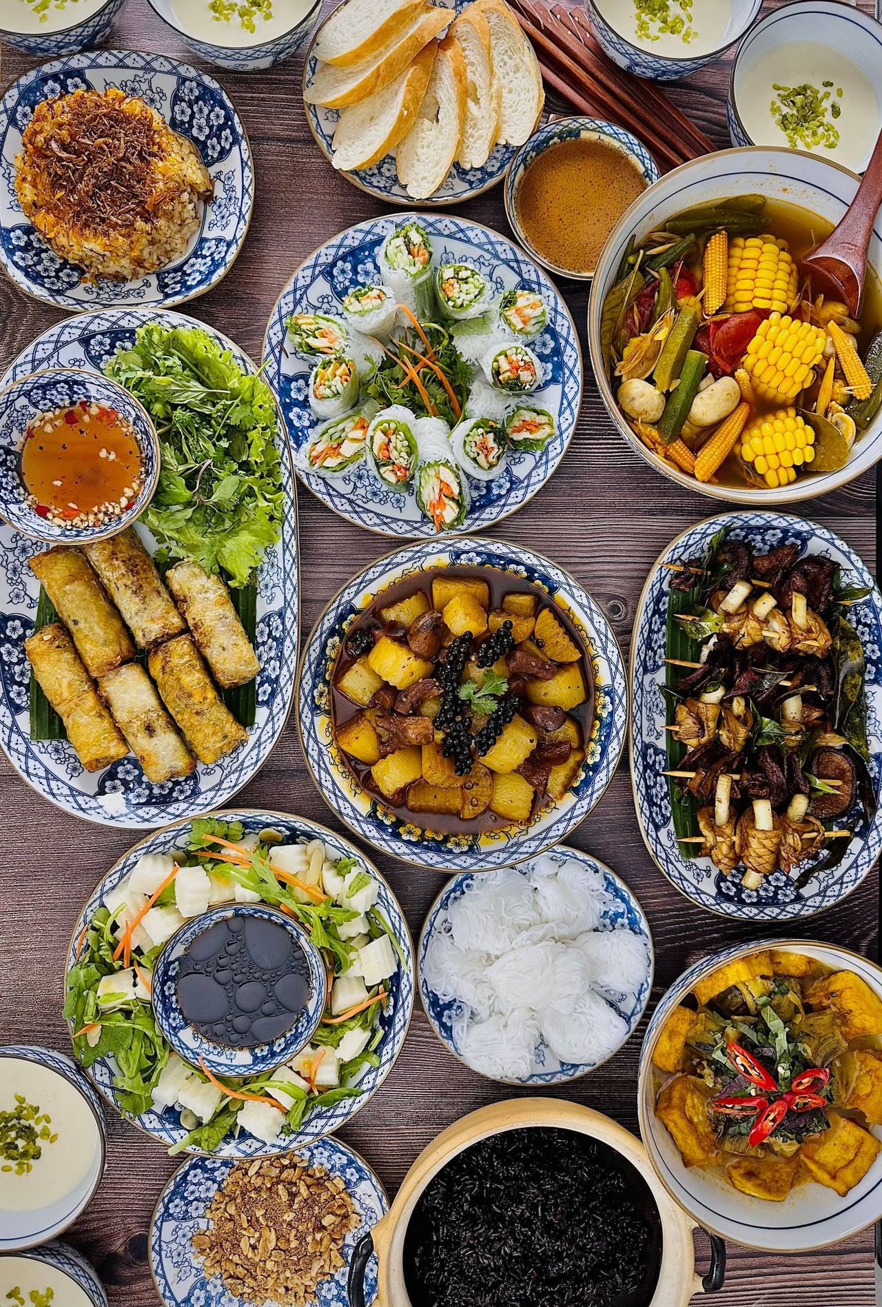 Cả mâm cơm nhìn từ trên: chả giò, gỏi cuốn, canh chua bắp, thịt kho, cơm và rau sống trên chén đĩa men lam