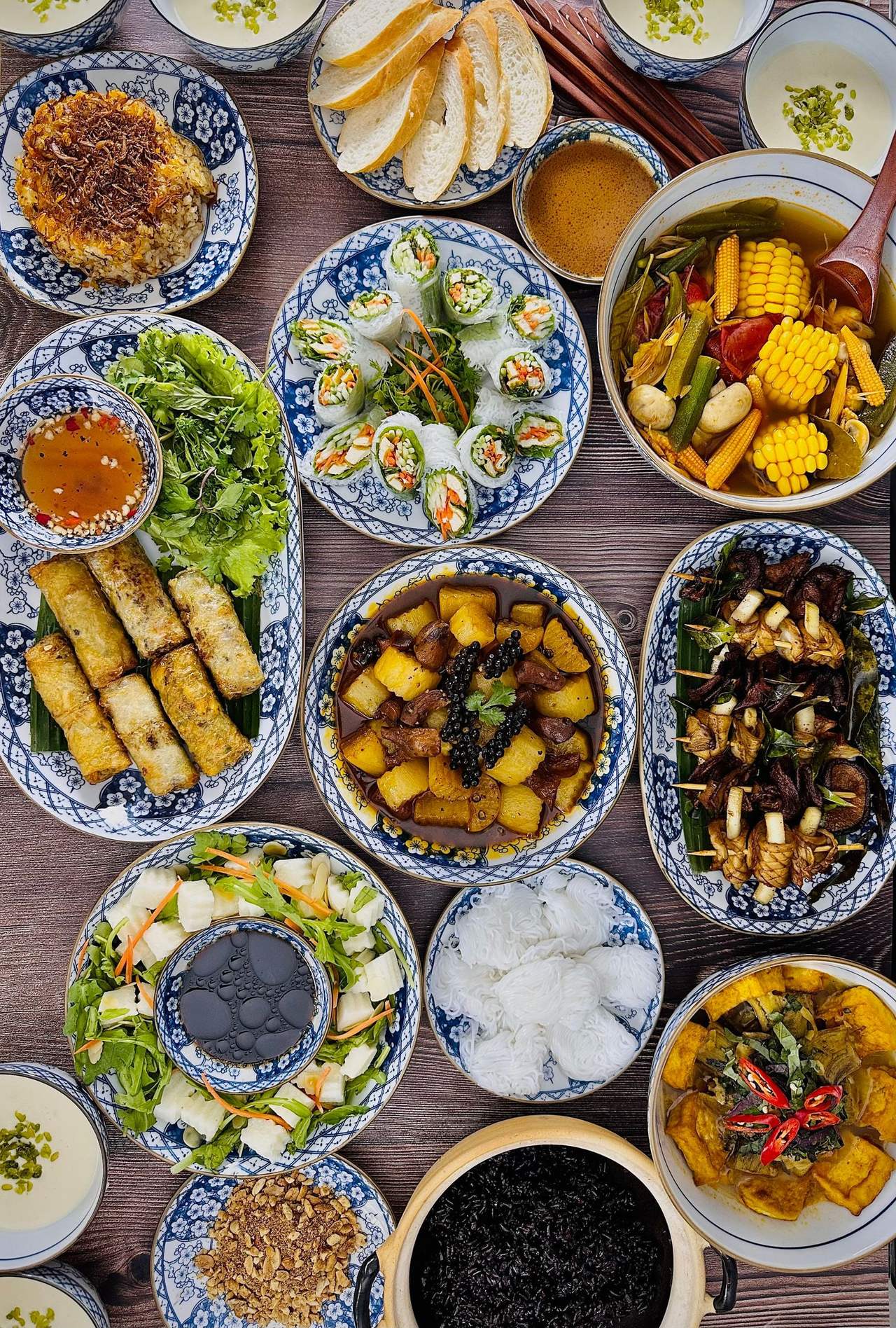 Cả mâm cơm nhìn từ trên: chả giò, gỏi cuốn, canh chua bắp, thịt kho, cơm và rau sống trên chén đĩa men lam