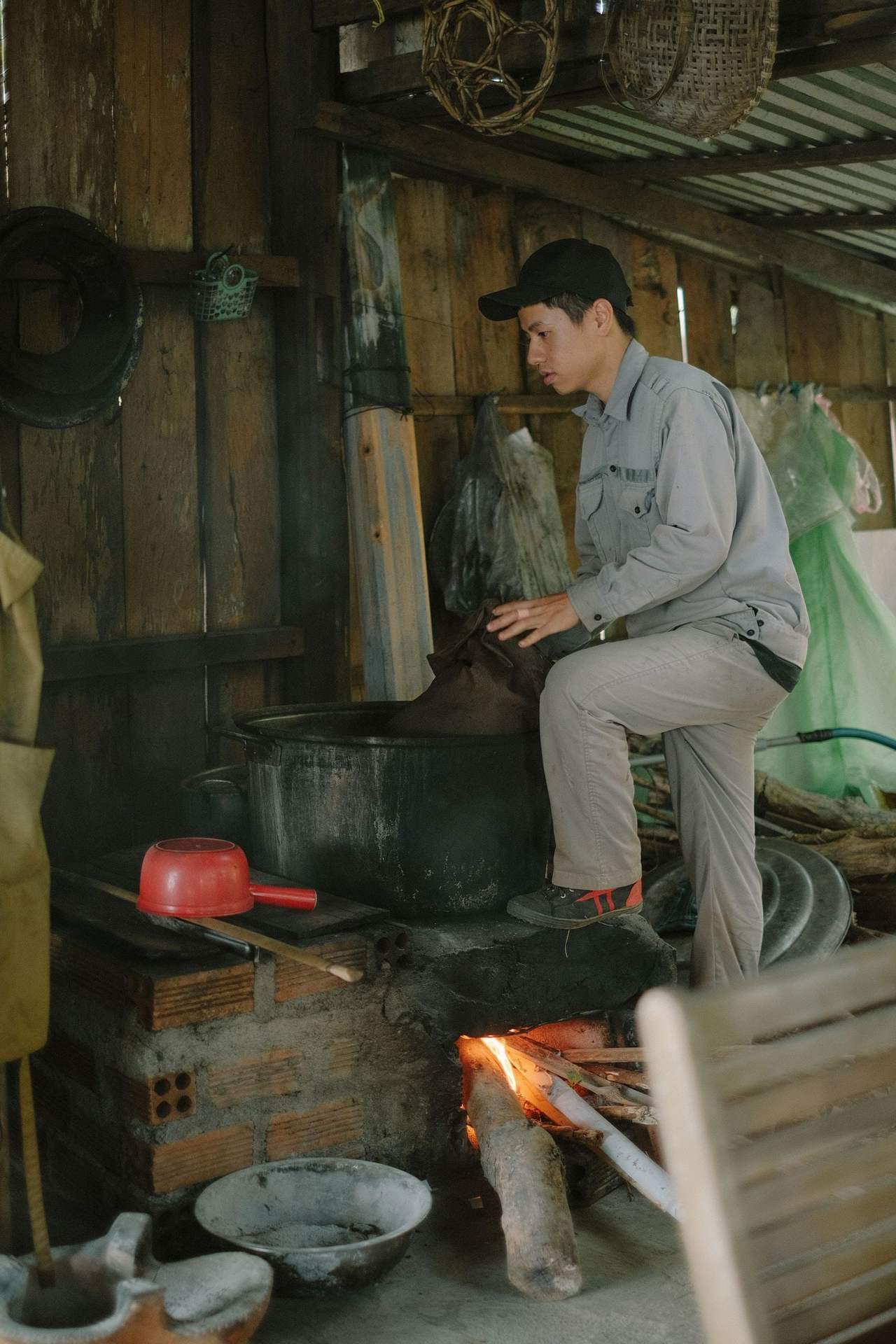 Người đàn ông ngồi bên bếp gạch đốt củi trong gian bếp gỗ, nhìn nghiêng khi canh lửa